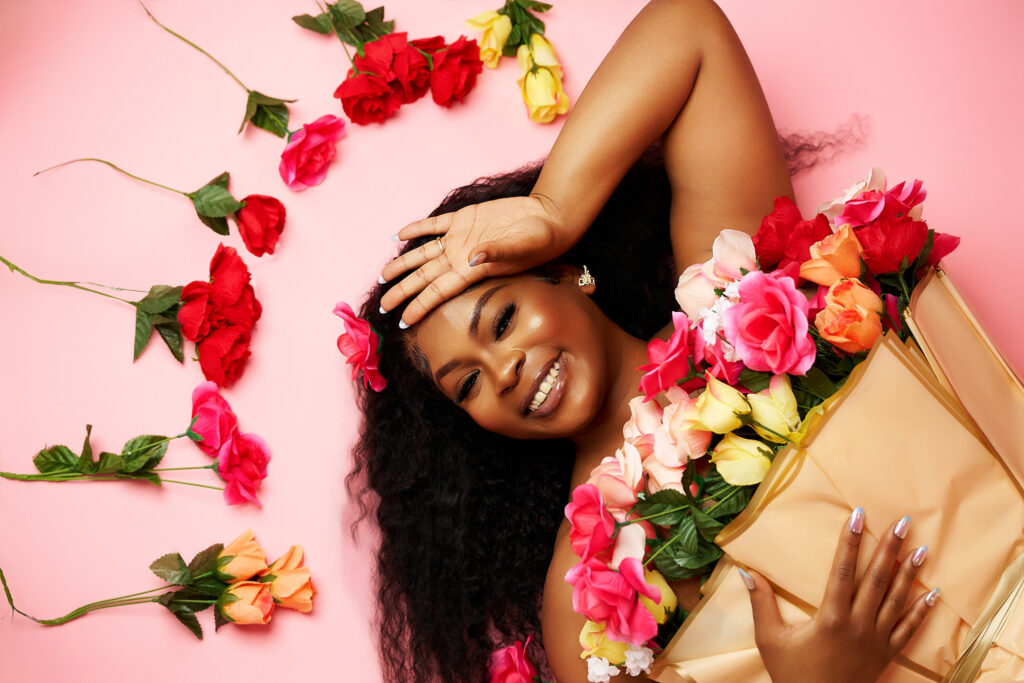 Overhead view of a joyful 27th birthday session in Houston, showing a smiling woman lying on a pink studio backdrop surrounded by scattered red, pink, and orange roses.