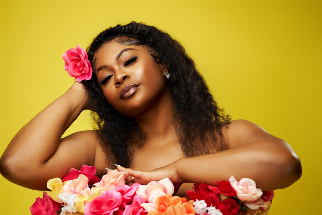 Close-up beauty portrait of a woman with glowing makeup and a pink rose in her hair, leaning gracefully against a bouquet of colorful flowers in a Houston photography studio with a yellow background.