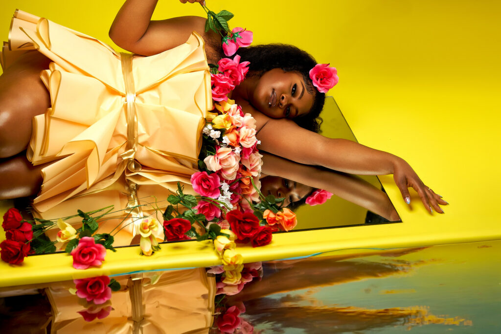 A creative Houston birthday portrait of a Black woman resting on a mirrored floor against a vibrant yellow studio backdrop, wearing a custom dress styled like a massive flower bouquet.