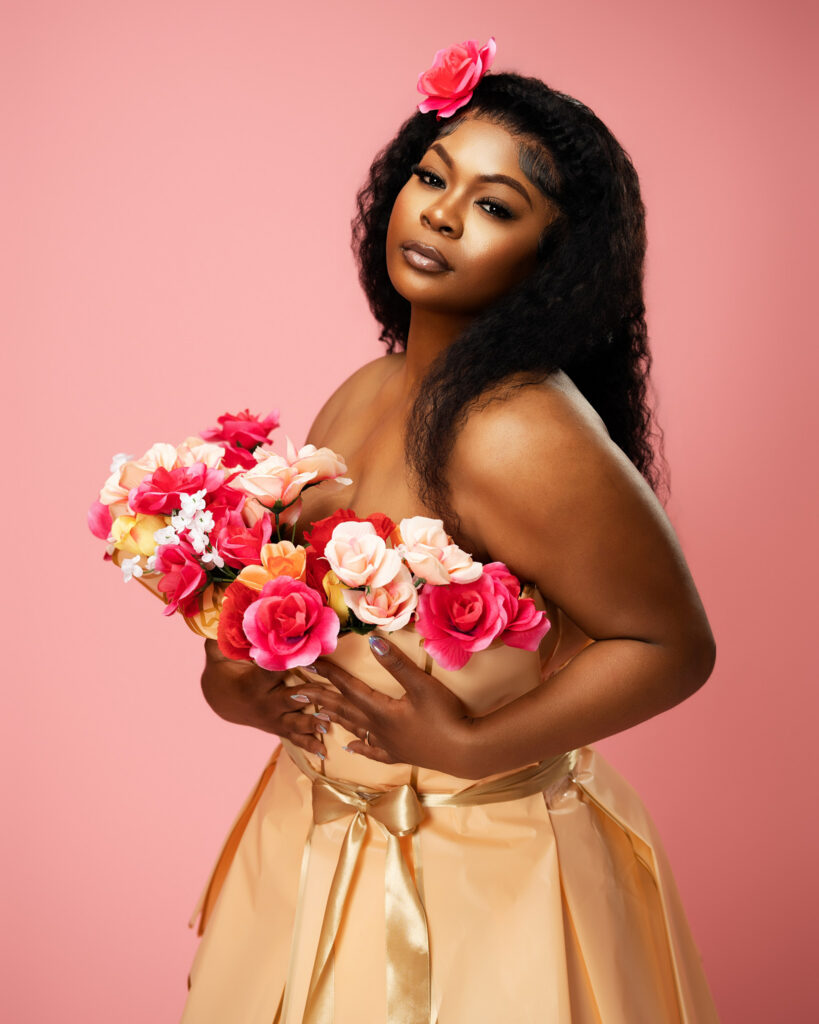 A Houston milestone birthday portrait of a woman standing gracefully against a seamless pink backdrop, modeling a unique artistic dress crafted from peach wrapping paper and faux roses